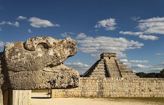 Een stenen beeld van het hoofd van Kukulcán. Op de achtergrond is El Castillo te zien, met de tempel bovenop.
